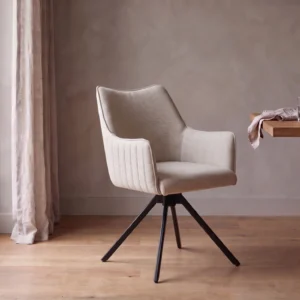 Eetkamerstoel Nicole in nude beige stof met armleuningen en zwarte poten aan een houten eettafel.